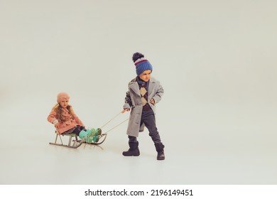 Portrait Of Boy And Girl, Children In Winter Clothes Sledding Isolated Over Grey Studio Background. Winter Holidays. Concept Of Childhood, Friendship, Fun, Lifestyle, Fashion, Retro Style