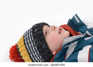 Portrait Of Boy Lying In Snow And Looking Up Into Sky. Child Is Wearing Multi-colored Knitted Hat. Close Up.