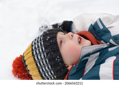 Portrait Of Boy Lying In Snow And Looking Up Into Sky. Child Is Wearing Multi-colored Knitted Hat. Close Up.