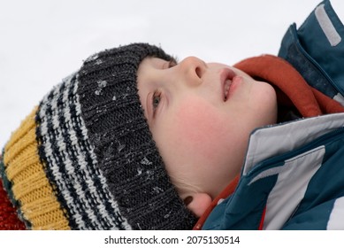 Portrait Of Boy Lying In Snow And Looking Up Into Sky. Child Is Wearing Multi-colored Knitted Hat. Close Up.