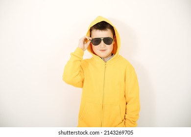 Portrait Of A Boy With Sunglasses  On White Background. Cool Kid Wearing Yellow Hoodie And Black Sunglasses. Child With A Hood On His Head