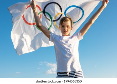 Portrait Of Boy Waving Flag The Olympic Games Outdoors Over Cloudy Sunset Sky. Children Sports Fan. Summer Olympic Games Concept. 08.06.2021, Barnaul Russia.