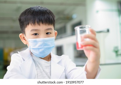 Portrait of a boy wearing hygiene mask and lab coat holding beaker which have pink solution in the laboratory. Kid looking forward and fun on experiment. Education, knowledge and learning concept