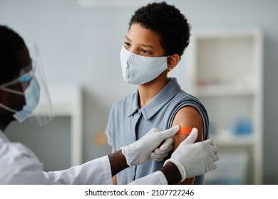 Portrait of brave African-American girl getting vaccinated in child vaccination clinic with doctor putting sterile plaster on shoulder
