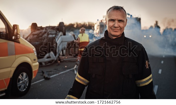 Portrait of the Brave\
Firefighter Smiling on Camera. In the Background Courageous Heroes\
Paramedics and Firemen Rescue Team Fight Fire, Smoke and Save\
People’s Lives