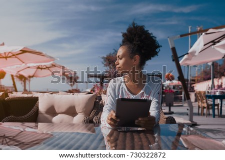 Portrait of Brazilian serious lady sitting in nearshore cafe, waiting her friend and holding digital pad; charming young black woman sitting with digital tablet in street bar of resort city near sea