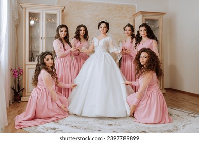 Portrait of the bride with her friends in the room. A brunette bride in a long white dress and her friends in pink dresses are smiling and happy. Young girls. - Powered by Shutterstock