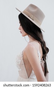 Portrait Of The Bride. A Young Attractive Brunette Woman With Long Black Hair In Braids. White Wedding Dress And Beige Hat. Rustic Style, A Bouquet Of Wheat Spikes. Back View.