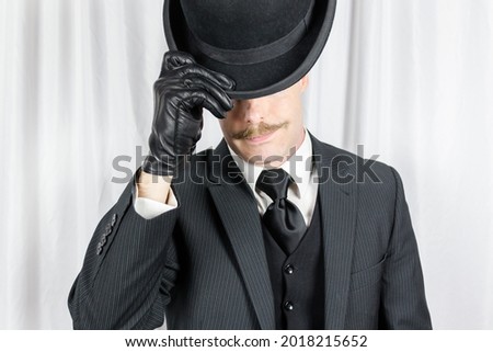 Portrait of British Gentleman in Dark Suit and Leather Gloves Politely Doffing Bowler Hat. Suave Spy Hero with Jaunty Moustache.