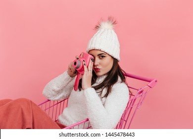 Portrait of brown eyed woman in white hat and sweater taking picture on instax while sitting in supermarket trolley on pink background