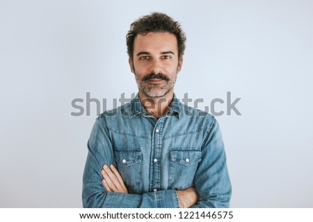 Portrait of brown, smiling, handsome man with mustache in jeans shirt standing with crossed arms. Gray background.