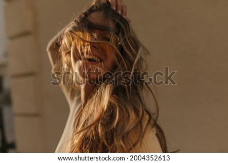 Portrait of brown-haired lady having fun ruffling her hair close-up against brown wall. On face are translucent yellow round glasses.