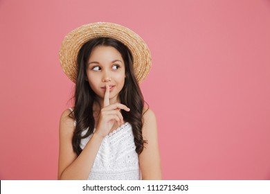 Portrait of brunette cute girl 8-10 wearing white dress and straw hat looking aside at copyspace with holding finger at lips isolated over pink background