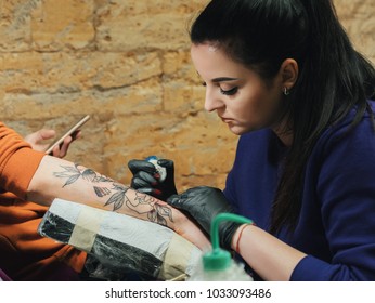 Portrait of brunette female tattooer in violet clothing making tattoo on the man's arm of her client with gun in studio. Young business woman in workshop.