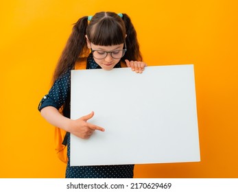 Portrait Of A Brunette Girl Child With Glasses Schoolgirl Girl Child Kid Pointing Finger On White Poster Banner Isolated On Yellow Color Background, Advertising, Billboard, Mockup