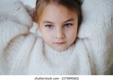 Portrait Of A Brunette Little Girl In White Fluffy Knitted Sweater Putting Her Hair In A Pony Tail. At Home, In Front Of A Curtain. Close Up. Looking Nowhere.