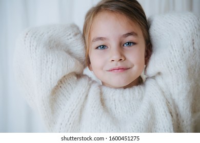 Portrait Of A Brunette Little Girl In White Fluffy Knitted Sweater Putting Her Hair In A Pony Tail. At Home, In Front Of A Curtain. She's Looking At Camera. Close Up.