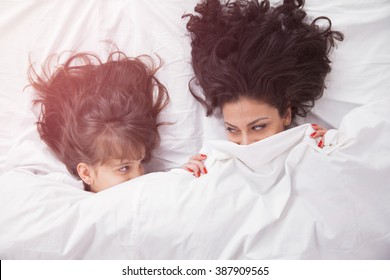 Portrait Of Brunette Long Hair Mother And Daughter Under The Duvet Together In Soft Morning Light On White Linen Bed. Concept Of Happy Family Living, Relaxation, Comfort, Fun. Top View.