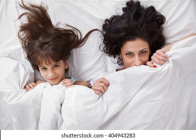 Portrait Of Brunette Long Hair Mother And Daughter Under The Duvet Together In Soft Morning Light On White Linen Bed. Concept Of Happy Family Living, Relaxation, Comfort, Fun. Top View.