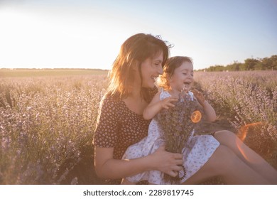 Portrait of brunette mother with little daughter sitting in purple lavender field. Young woman in rural dress lovingly embraces, kisses girl. The concept of allergy, travel, single parent. Copy space