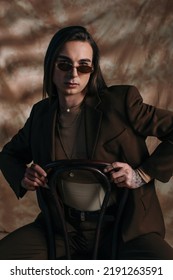 Portrait Of Brunette Nonbinary Person In Sunglasses And Jacket Sitting On Chair On Abstract Brown Background