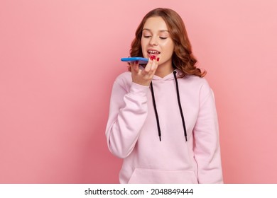 Portrait Of Brunette Teen Girl In Hoodie Talking To Smartphone, Using Virtual Assistant, Ai Intelligent Technology, Voice Application, Audio Message. Indoor Studio Shot Isolated On Pink Background