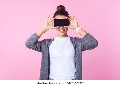 Portrait Of Brunette Teenage Girl With Bun Hairstyle In Casual Clothes Smiling And Covering Eyes With Cell Phone, Hiding Face To Be Anonymous In Internet. Indoor Studio Shot Isolated, Pink Background