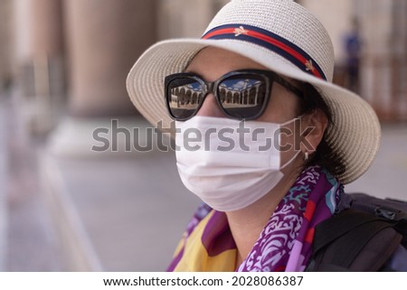 Portrait of a brunette woman in a protective mask on her face, in sunglasses and in a white hat on her head. Reflection in sunglasses building with arches and sky.