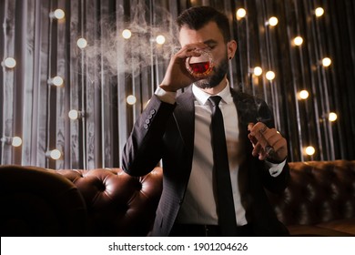 Portrait Of A Brutal Beard Man With Tattoo Smoking Cigar In Hot Elegant Suit  Drinking Whiskey Alcohol In Loft Apartment . Couple Love Story Concept