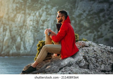 Portrait of a brutal bearded man in a red jacket by the sea. Male portrait on the background of a seascape. A traveler on the background of the sea coast. A man in a jacket at sunset