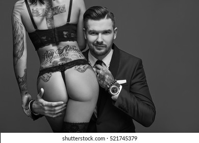 Portrait of a brutal man in elegant suit touches sexy girl with a tattoo in lingerie on gray background