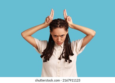 Portrait Of Bully Woman With Black Dreadlocks Looking Threatening And Showing Bull Horn Sign, Holding Fingers On Her Head, Conflicting Person. Indoor Studio Shot Isolated On Blue Background.