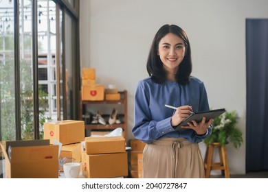 Portrait business Asian woman smiling look at camera and use tablet checking information on parcel shipping box before send to customer. Entrepreneur small business working at home.