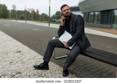 Portrait Of A Business Man With A Laptop In His Hands Talking On A Mobile Phone Next To An Office Building, Entrepreneurship Development And Business Planning Concept