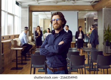 Portrait Of Business Teacher And Team Coach. Happy Handsome Bearded Young Man In Blue Shirt And Eyeglasses Standing In Office After Corporate Training Class, With Group Of Employees In Background