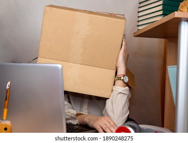 Portrait Of Business Woman With Box Over Head. Carton Box Contains Sign Thinking Inside The Box. She Is Sitting At Chaotic Desk, With Loth Of Things On It. Home Office.