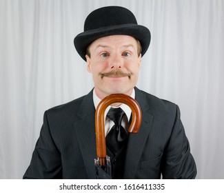 Portrait of Businessman in Dark Suit and Bowler Hat Smiling With Chin Resting on Umbrella. Classic and Eccentric British Gentleman Stereotype. Quirky and Cute Character. Sidekick to Action Hero