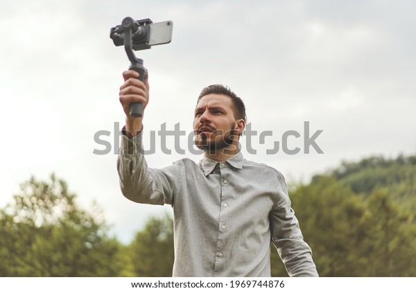 Portrait of businessman recording video presentation at\
smartphone with steadycam. Focused bearded man standing outside,\
holding steadycam while recording video presentation about new\
object 