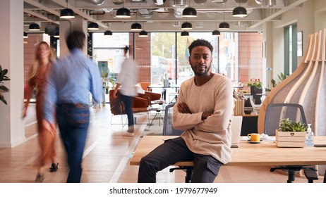 Portrait Of Businessman Sitting On Desk In Busy Multi-Cultural Office With Motion Blurred Colleagues
