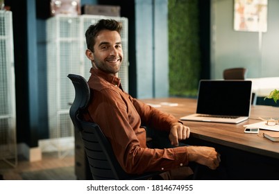 Portrait Of A Businessman Smiling While Working Late On A Laptop At His Desk In A Quiet Office