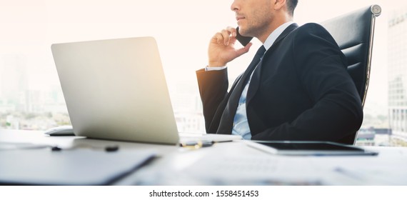 Portrait of Businessman talking business on phone at the office. Handsome man in black suit meeting via smartphone while working with laptop computer near window. Banner.