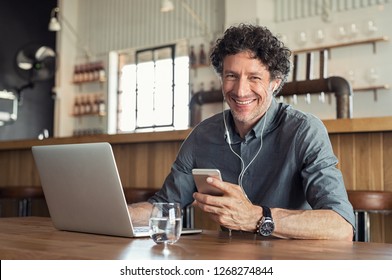 Portrait of businessman using smartphone with earphones and laptop. Smiling business man sitting in restaurant listening to music. Happy man enjoying free wireless internet connection at coffee shop.