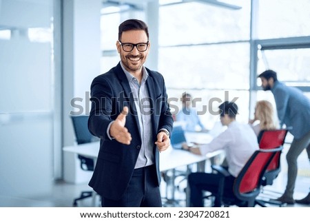 Portrait of businessman welcoming new employee to his business team and company, giving hand forward in modern office with colleagues in the background.