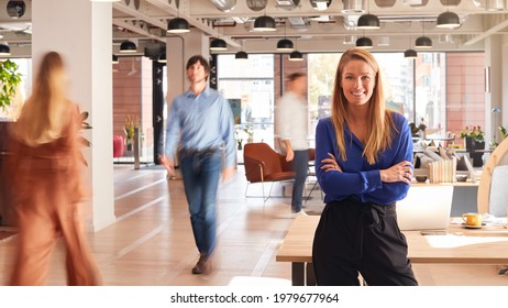 Portrait Of Businesswoman By Desk In Busy Multi-Cultural Office With Motion Blurred Colleagues