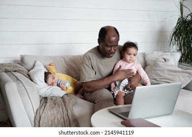 Portrait Of Busy African American Dad Sitting On Sofa In Living Room Taking Care Of His Twin Babies And Working In Internet