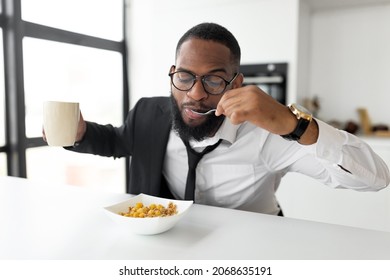 Portrait of busy African American man in glasses having breakfast in kitchen at home, wearing suit holding cup drinking coffee while eating cereal, rushing to office, hurry to work in the morning - Powered by Shutterstock