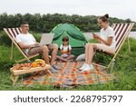 Portrait of busy family, mother, father and their daughter spending time near the river, sitting on deck-chairs near the tent, husband doing his online work, wife using smart phone, kid eating fruit.