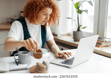 Portrait of busy female project manager working from home at kitchen table eating cookies unconsciously taking one by one from transparent jar, scrolling on laptop, looking at screen attentively - Powered by Shutterstock