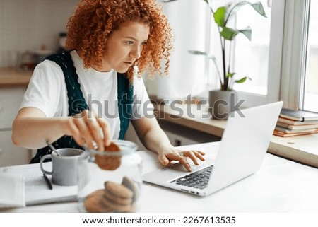 Portrait of busy female project manager working from home at kitchen table eating cookies unconsciously taking one by one from transparent jar, scrolling on laptop, looking at screen attentively