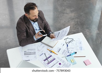 Portrait Of Busy Male Office Worker Makes Voice Call, Discuss Documents With Partner, Compare Result, Try To Predict Wastes For Next Month. Head Executive Officer Holds Balance Sheet And Mobile Phone